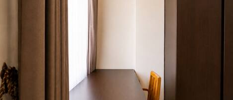 Una habitación estrecha con una sola silla de madera, una mesita y una ventana con cortinas.