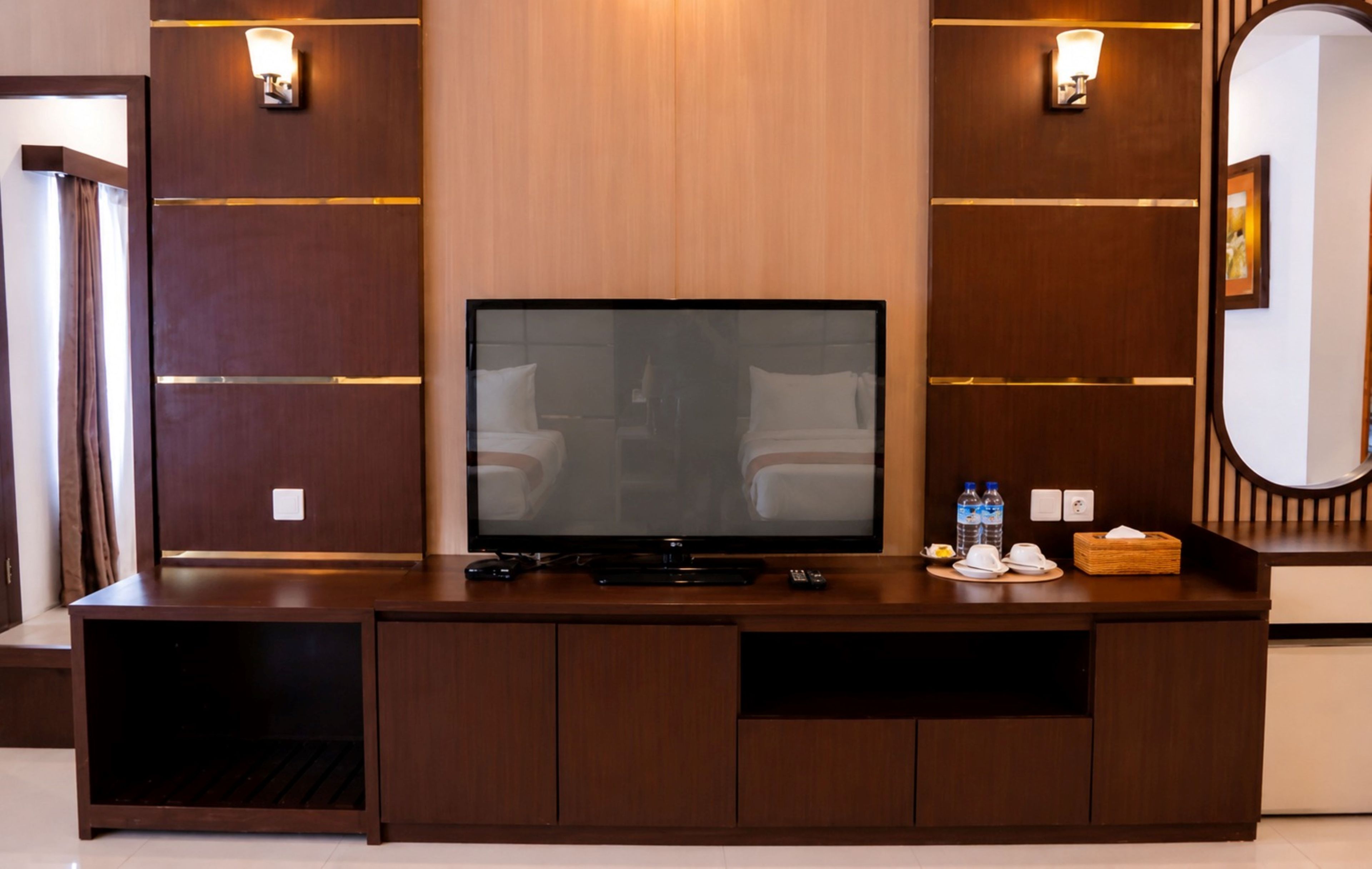 Habitación de hotel con un televisor de pantalla plana grande sobre un mueble de madera, un espejo y una mesita con una botella de agua y una caja de pañuelos.