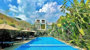 A rectangular swimming pool with clear blue water, surrounded by lounge chairs and umbrellas, with a building in the background. - Diva Lombok Resort (Senggigi)