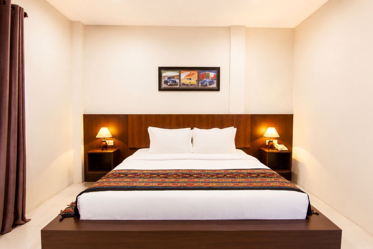 Quarto de hotel com uma cama grande, duas mesas de cabeceira com abajures, um quadro na parede e cortina marrom.