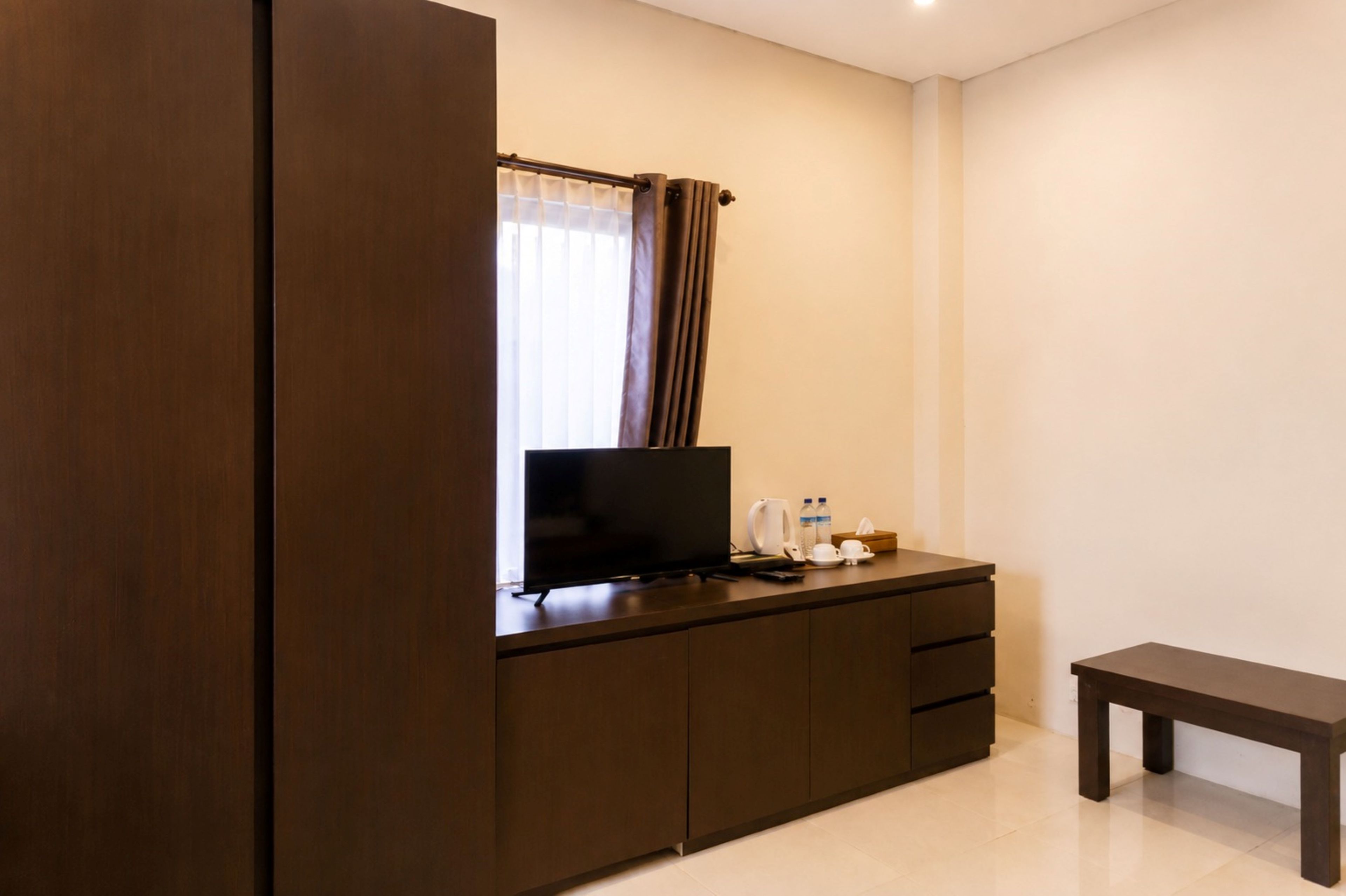 A hotel room with a dark wooden wardrobe, a flat-screen TV on a wooden cabinet, a small bedside table with a cup, a bottle, and a tissue box.