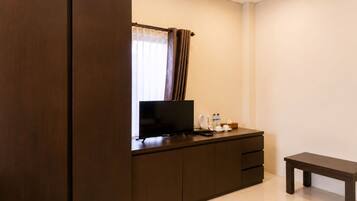 A hotel room with a dark wooden wardrobe, a flat-screen TV on a wooden cabinet, a small bedside table with a cup, a bottle, and a tissue box.