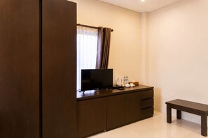 A hotel room with a dark wooden wardrobe, a flat-screen TV on a wooden cabinet, a small bedside table with a cup, a bottle, and a tissue box. - Diva Lombok Resort (Senggigi)