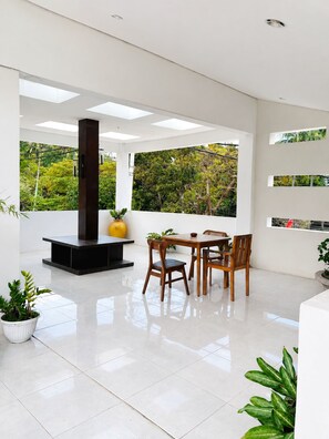 A modern interior with a dining area, a fireplace, and a view of greenery. - Diva Lombok Resort (Senggigi)