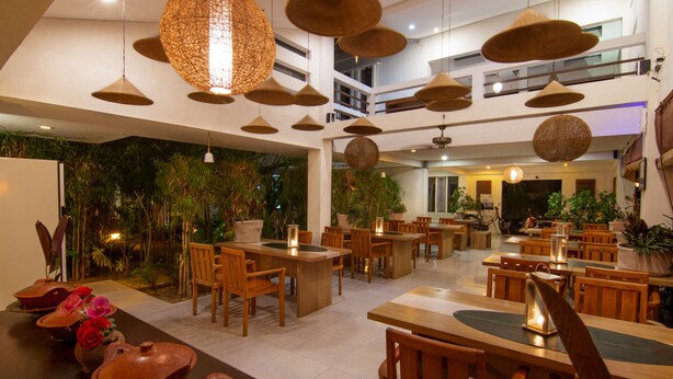A spacious indoor dining area with multiple wooden tables and chairs, surrounded by potted plants and hanging woven light fixtures. - Diva Lombok Resort (Senggigi)