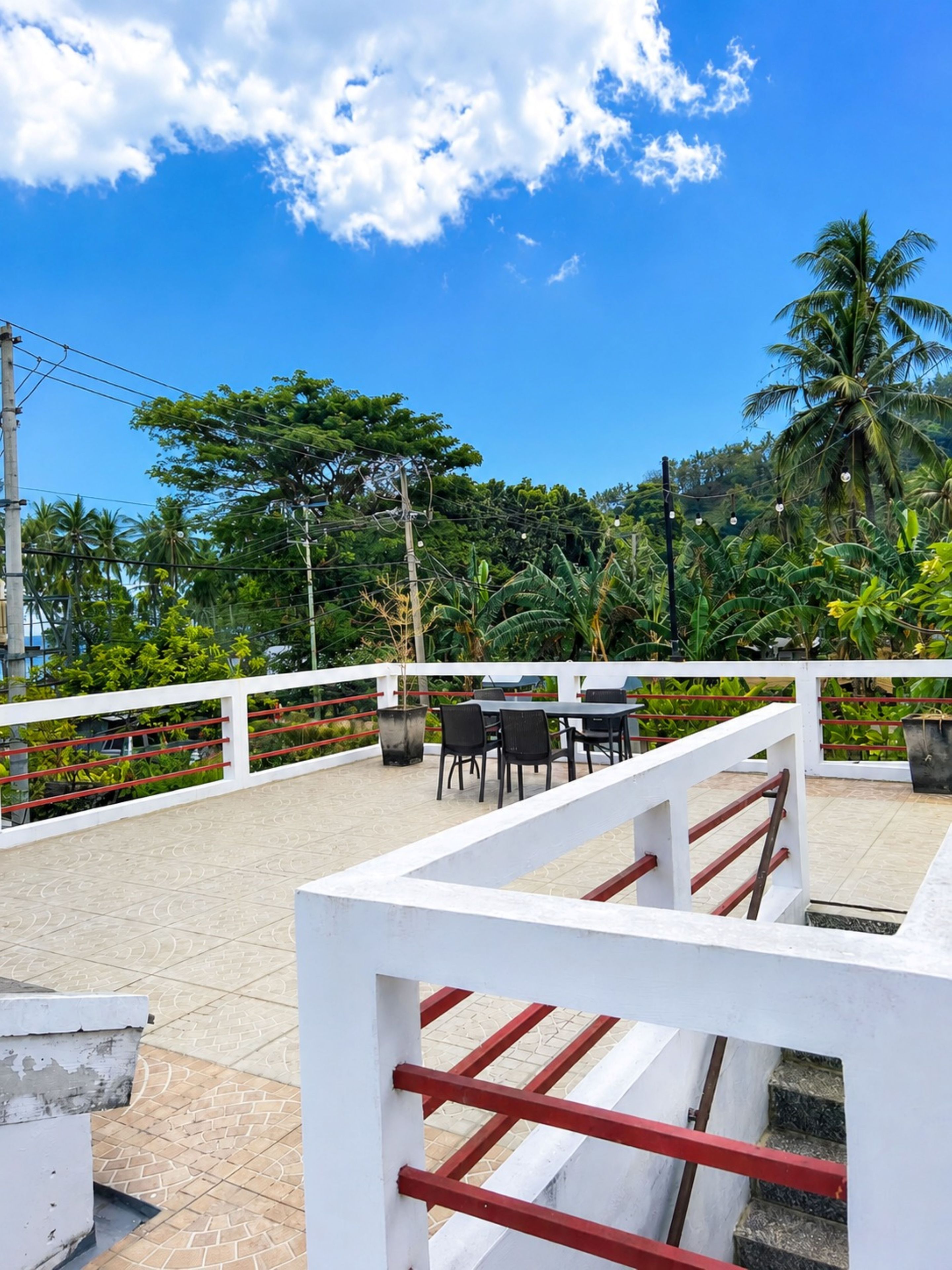 Une terrasse extérieure dotée d’une balustrade blanche, de tables et de chaises, entourée d’une végétation luxuriante et de palmiers.