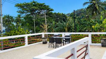 Une terrasse extérieure dotée d’une balustrade blanche, de tables et de chaises, entourée d’une végétation luxuriante et de palmiers.