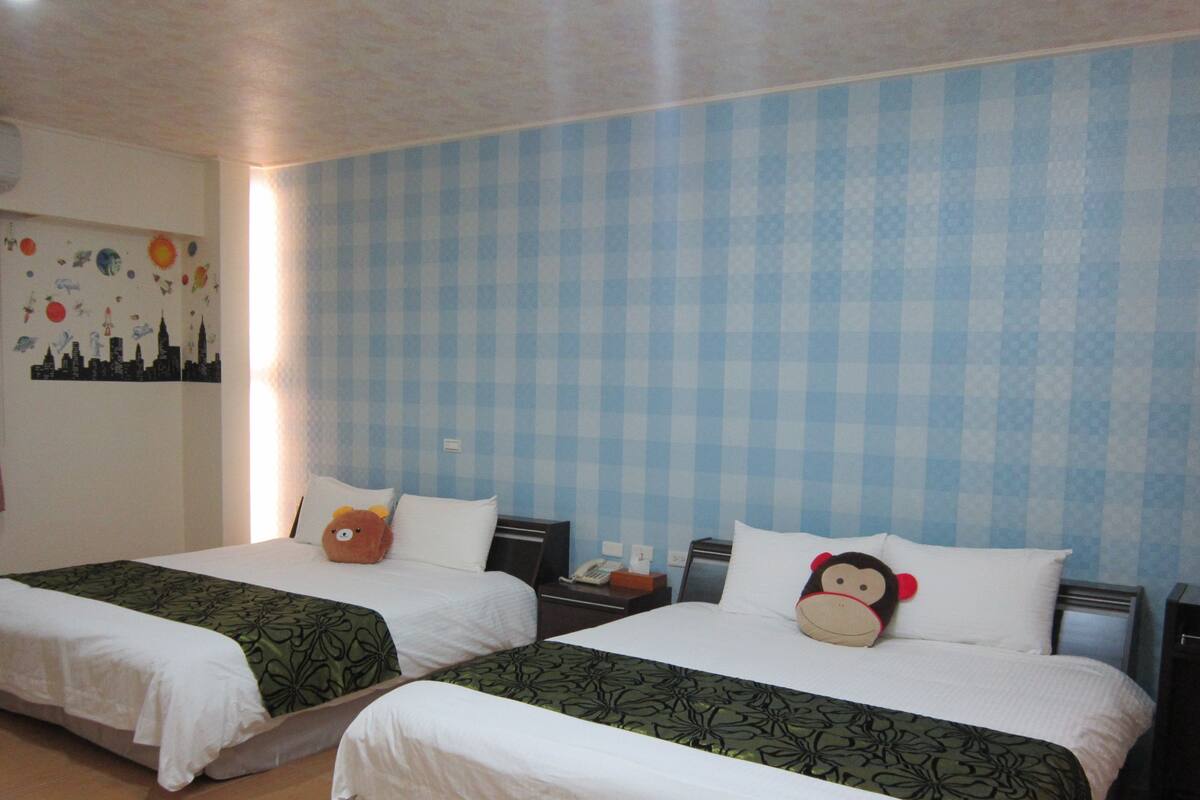 A hotel room with two beds, each with a green patterned bedspread and white pillows. There is a wall decoration with a cityscape and a wall clock.