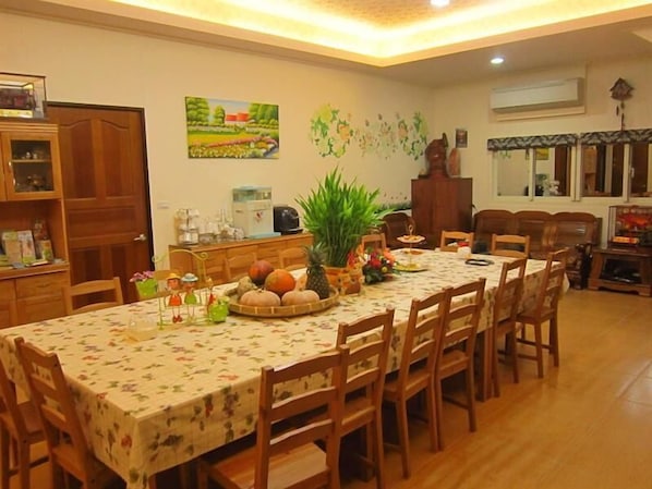 Un comedor con una mesa larga, sillas de madera y un mantel con motivos florales. Hay una planta en maceta, una cesta de frutas y un reloj en la pared.