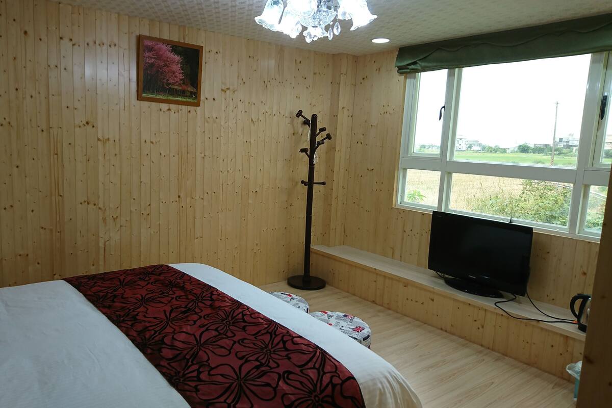 A wooden-paneled room with a bed, a television on a stand, and a window with a view of greenery.