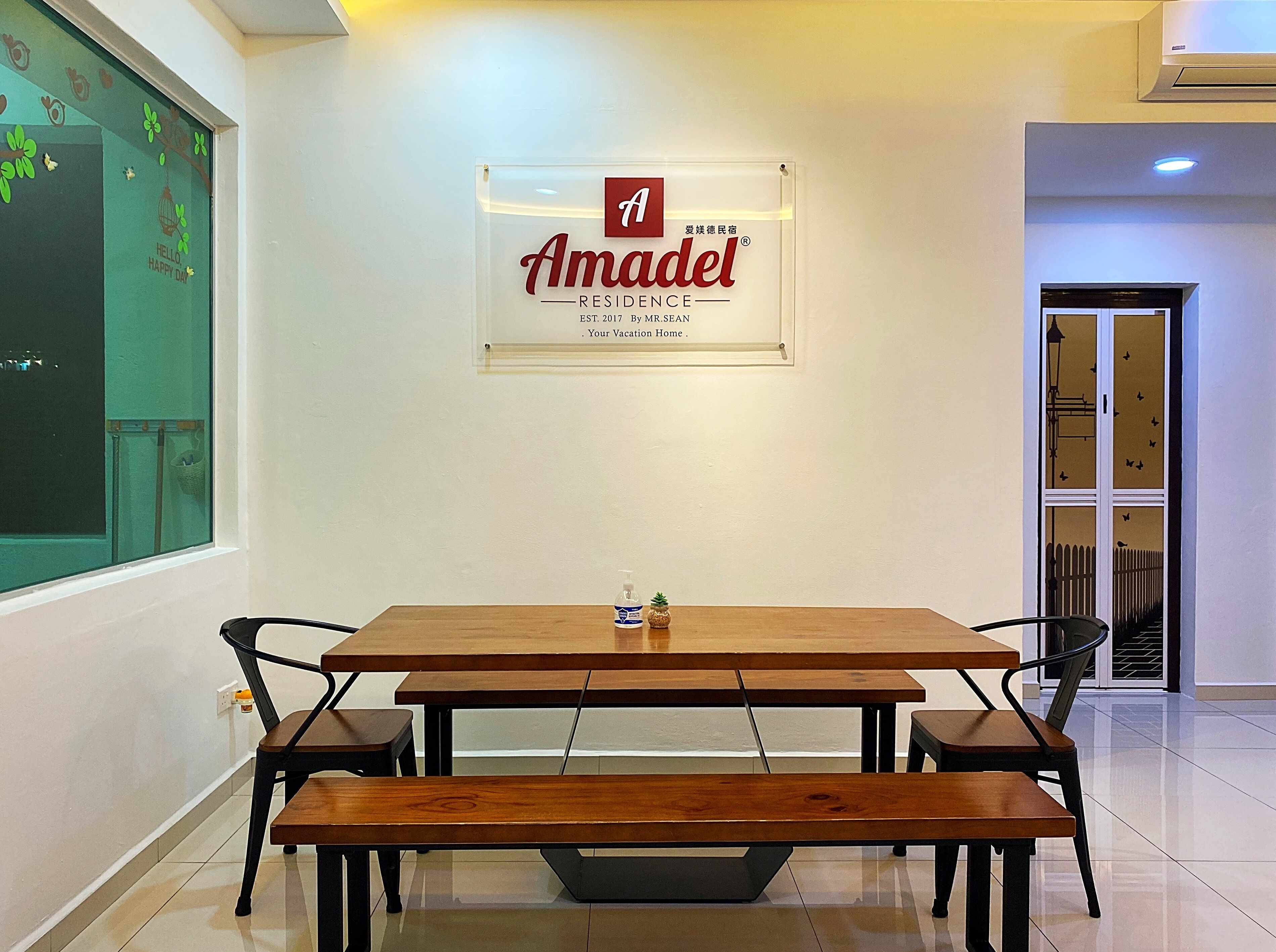 A conference room with a wooden table and benches, a sign for Amadel Residence, and a window with a view of the outside.