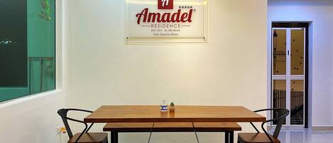 A conference room with a wooden table and benches, a sign for Amadel Residence, and a window with a view of the outside.