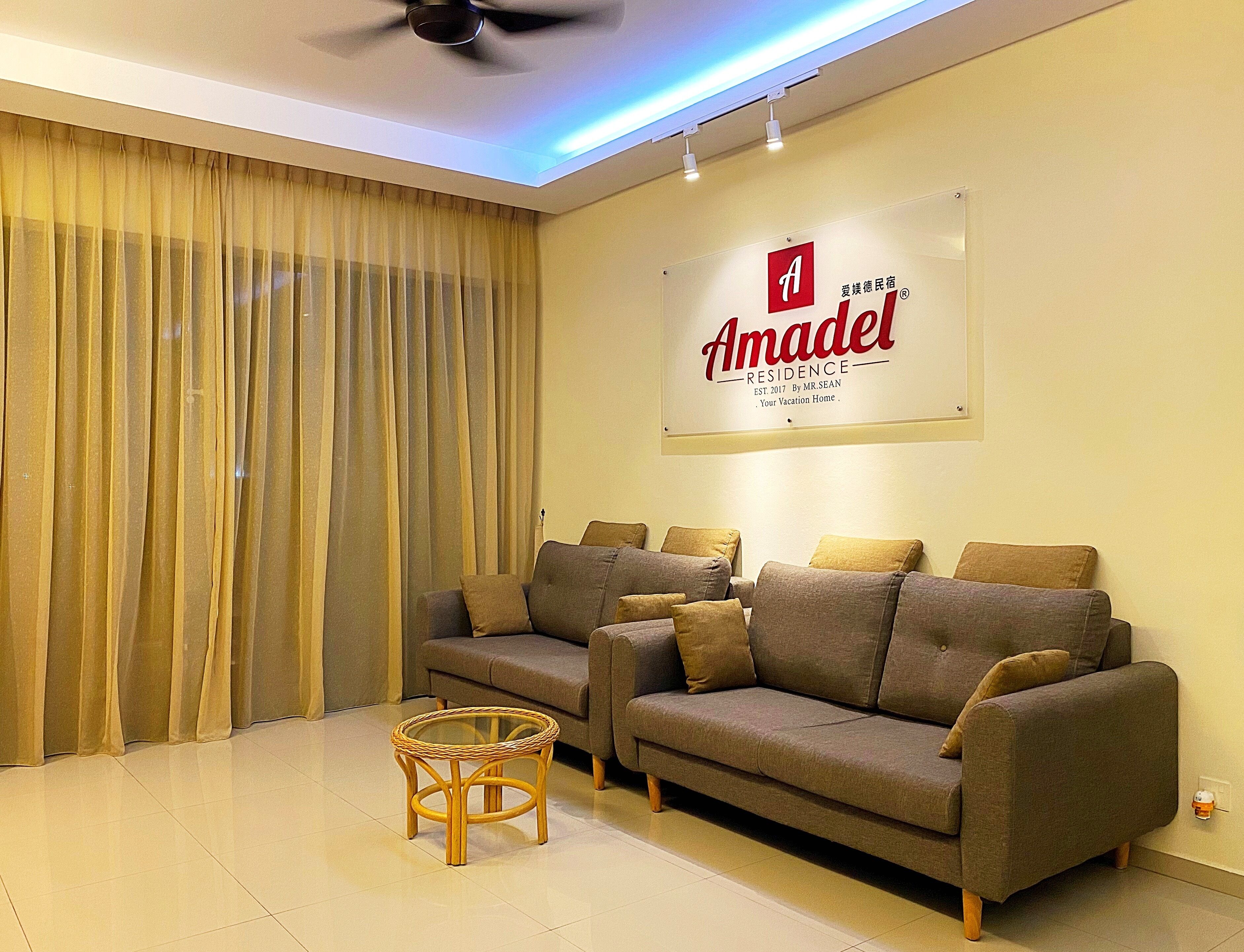 A living room with a sofa set, a glass coffee table, and a sign for Amadel Residence.