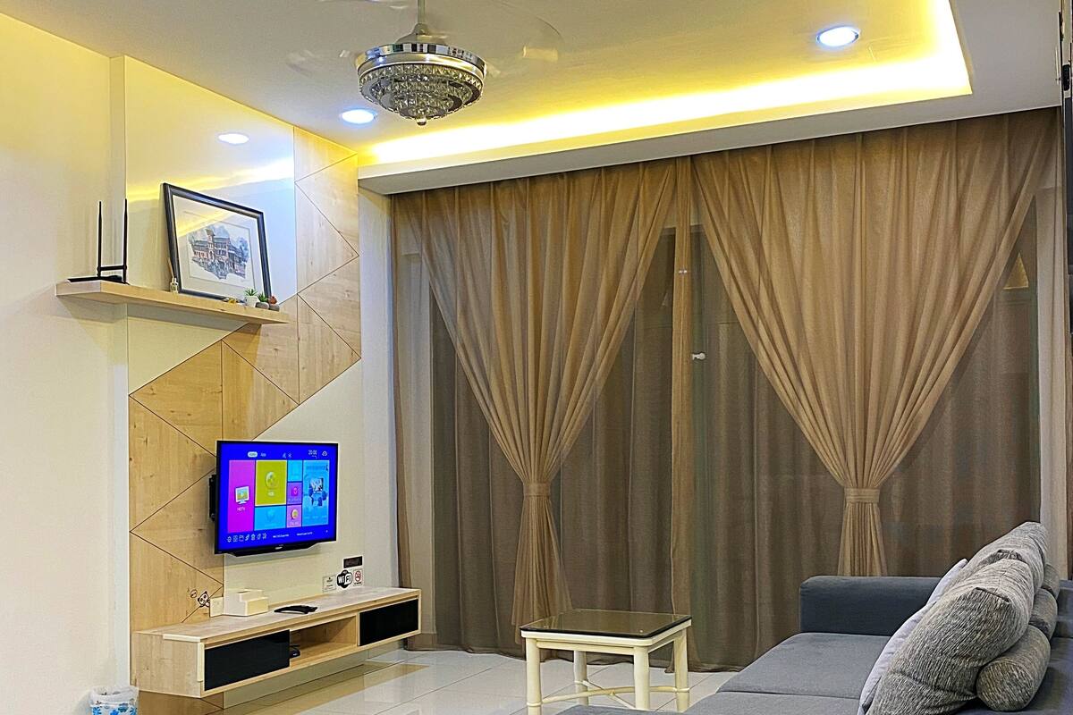 A modern living room with a grey sofa, a TV on a wooden stand, a ceiling fan, and a chandelier.
