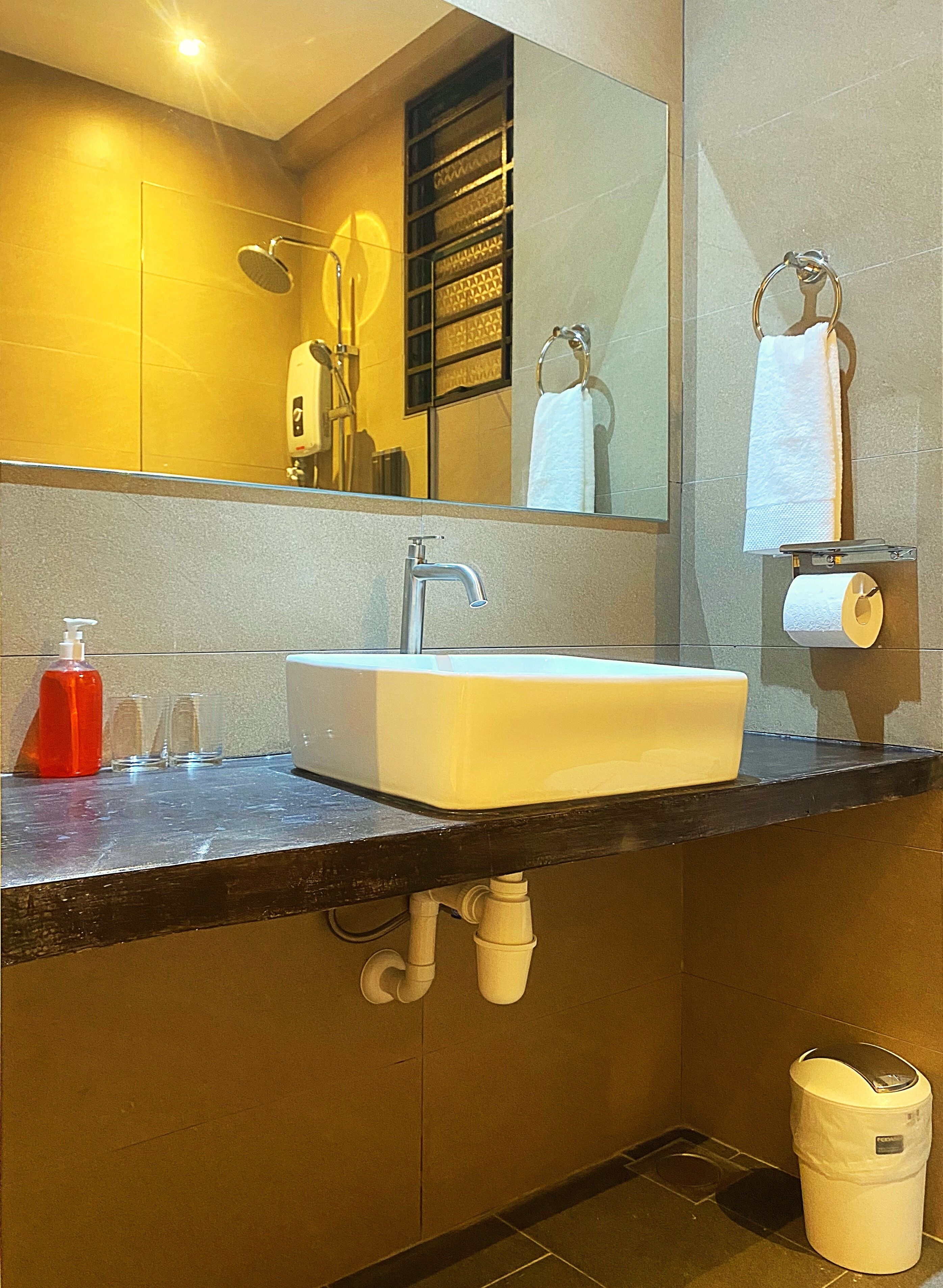 Un baño con lavamanos blanco, espejo, toallero, dispensador de jabón y cubo de basura.