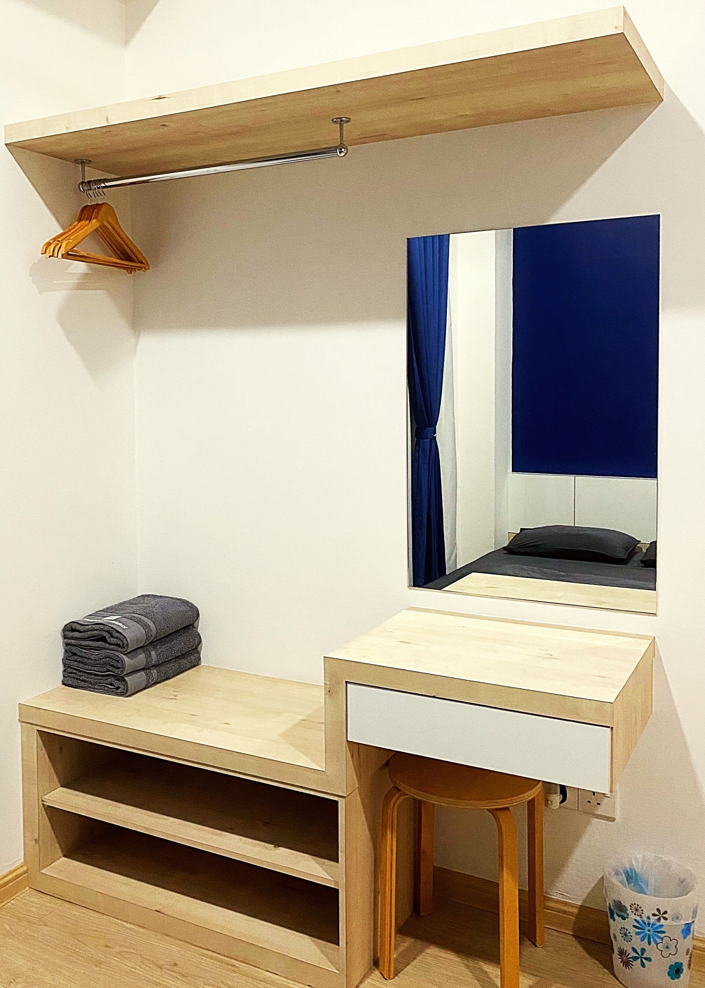 A compact bedroom with a wooden desk, a mirror, a hanging rack, and a small shelf.
