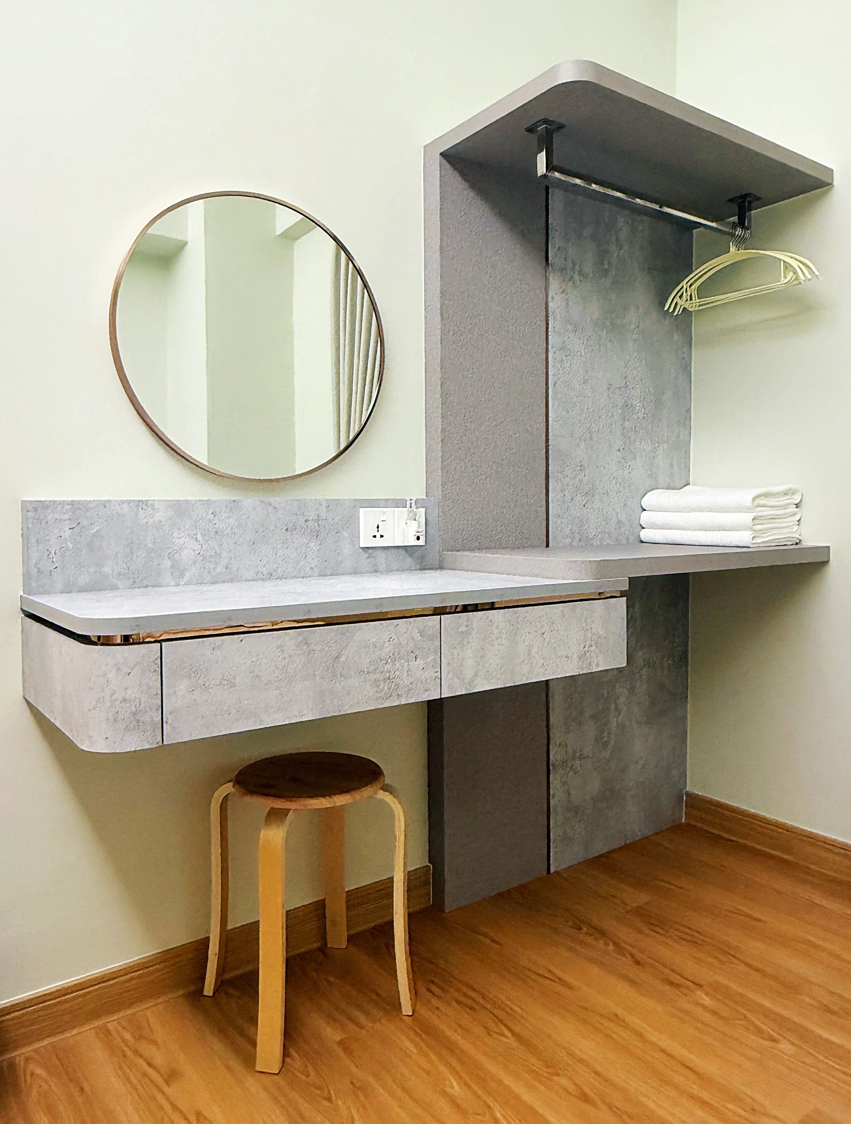 Un bagno con un piano di lavoro grigio, uno specchio rotondo, uno sgabello di legno e un porta asciugamani.