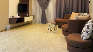 A modern living room with a flat-screen TV, a ceiling light fixture, a sofa, a small stool, and a large window with curtains. - Amadel Residence (Malacca City)