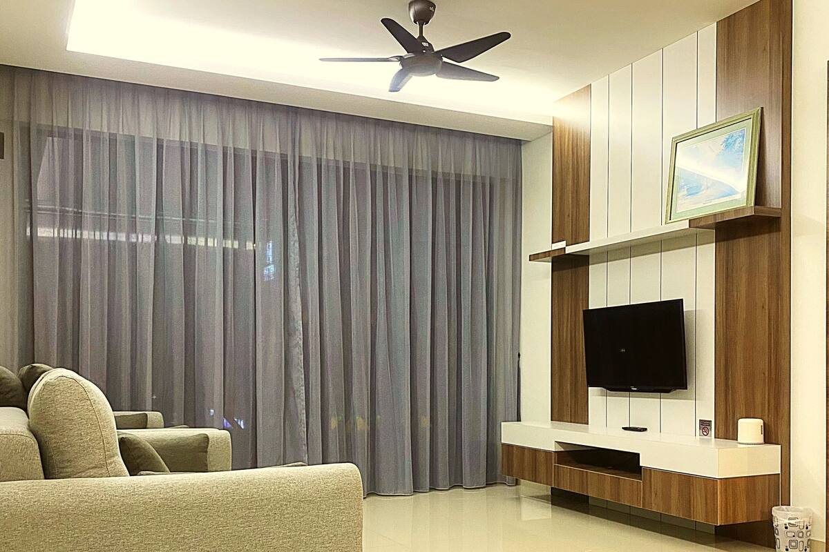 A modern living room with a sofa, a flat-screen TV mounted on a wooden panel wall, a ceiling fan, and sheer curtains.