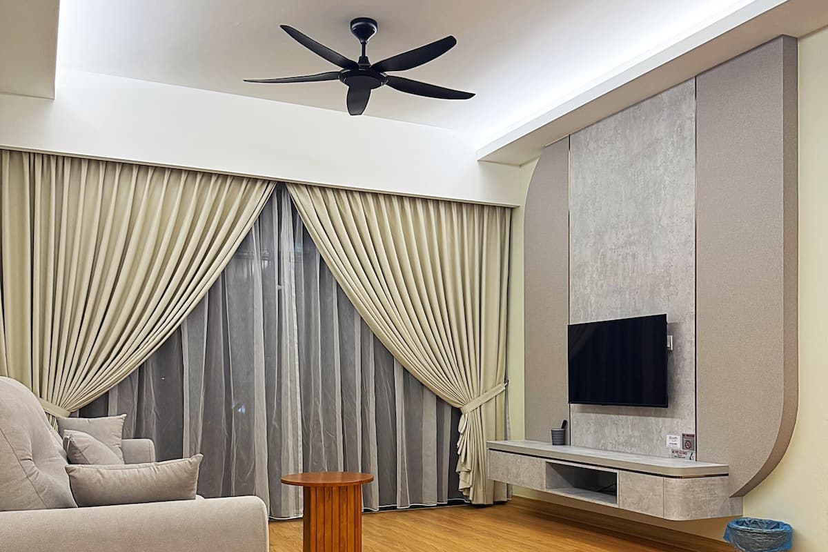 A modern living room with a sofa, a round wooden side table, a ceiling fan, a TV mounted on a wall, and a large window with curtains.