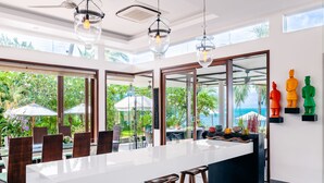 A modern kitchen with a large island, bar stools, and a view of a lush garden and ocean. - Lotus Samui (Koh Samui)