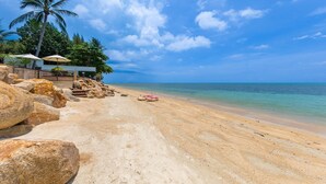 A beach with large rocks, a palm tree, and a building with a covered area. - Lotus Samui (Koh Samui)