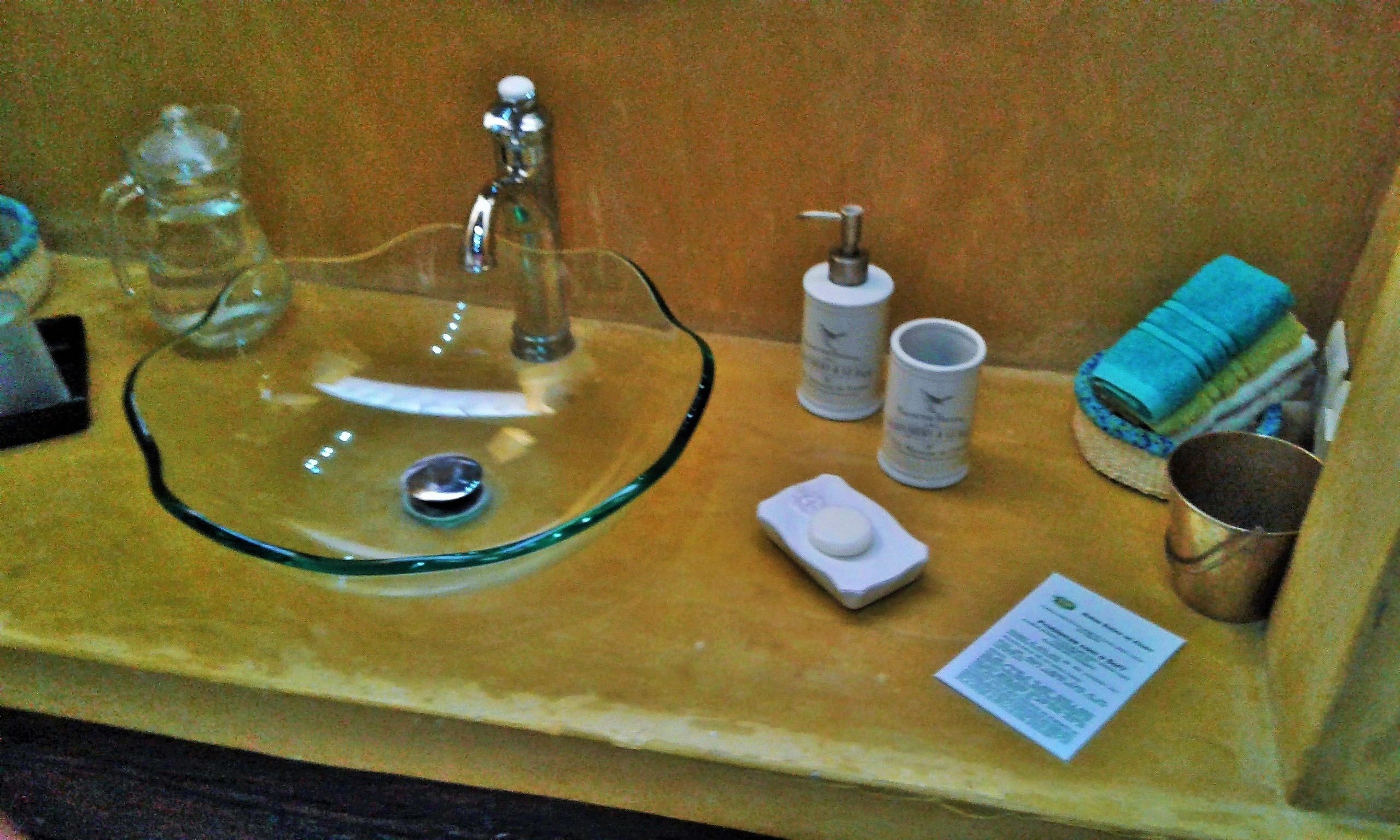 Un lavabo de vidrio con un grifo, un dispensador de jabón, un pequeño plato y una taza de metal sobre una encimera de madera.