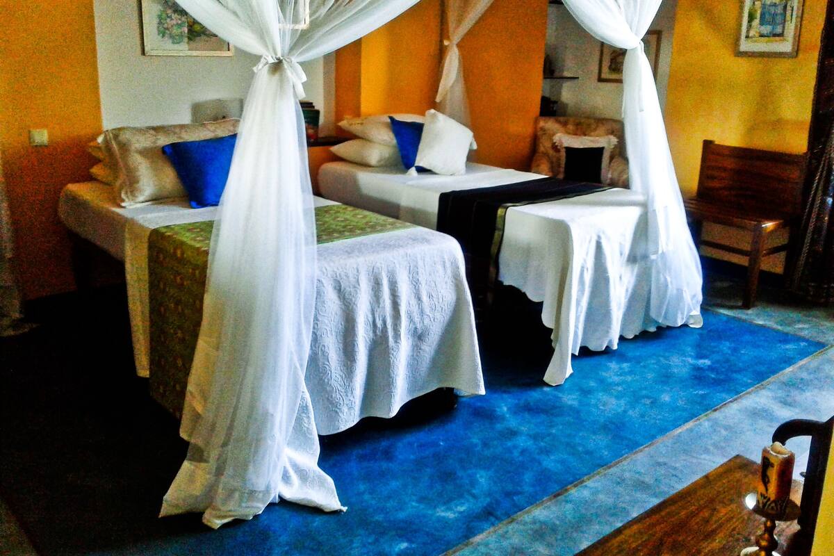 Une chambre avec deux lits, chacun recouvert de draps blancs et surmonté d’un baldaquin. On y trouve également une chaise en bois et une petite table.