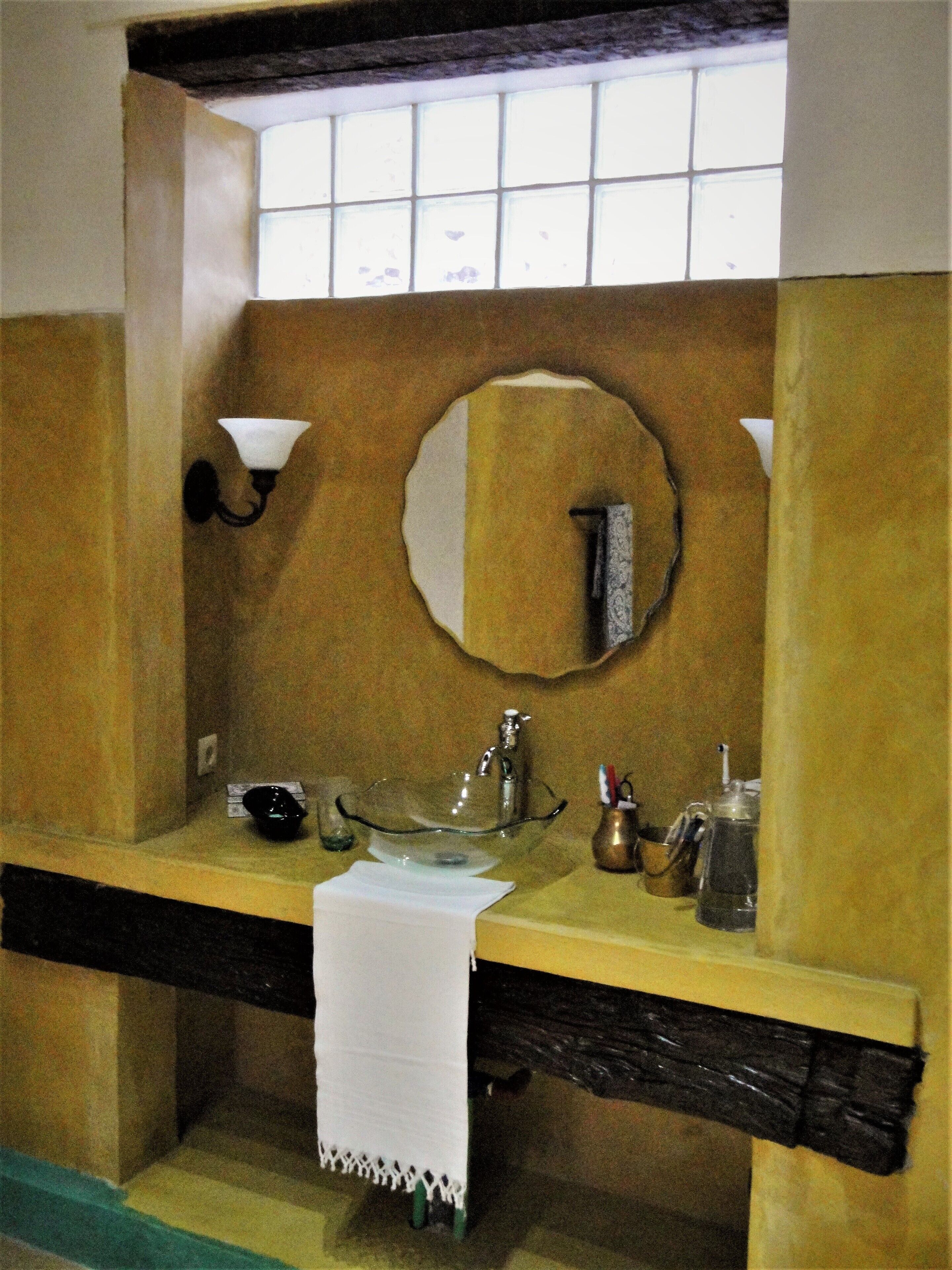 Un baño con tocador de madera, lavamanos de vidrio, espejo y toalla colgada en la pared.