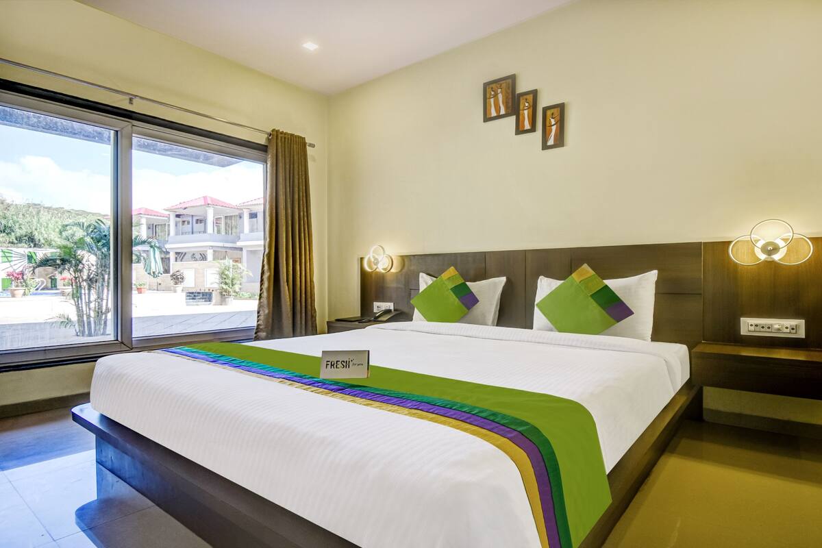 A hotel room with a large bed, a nightstand, a bedside table, a window with curtains, and wall-mounted lights.