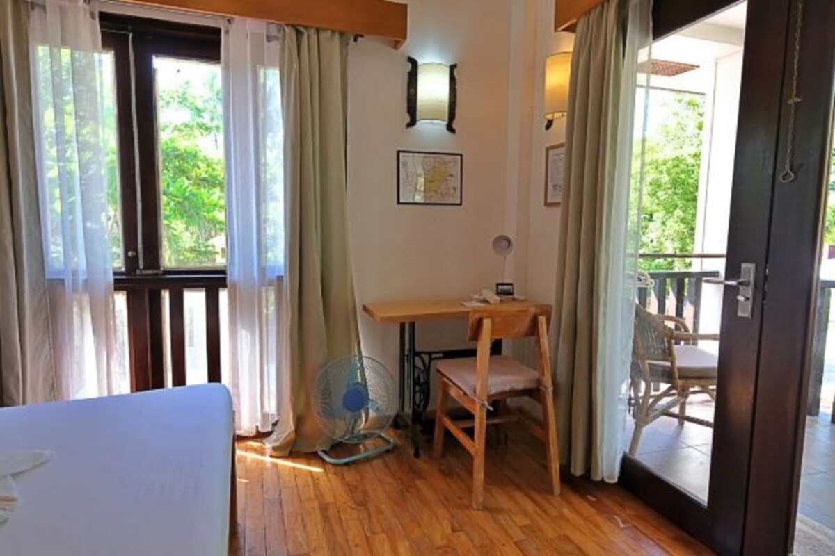 Una habitación con piso de madera, una cama, un escritorio con silla, un ventilador y una ventana con cortinas.