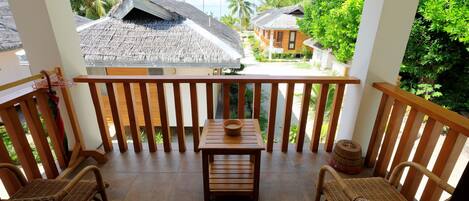 A balcony with wicker furniture and a small table, overlooking a tropical resort with thatched-roof buildings and palm trees.