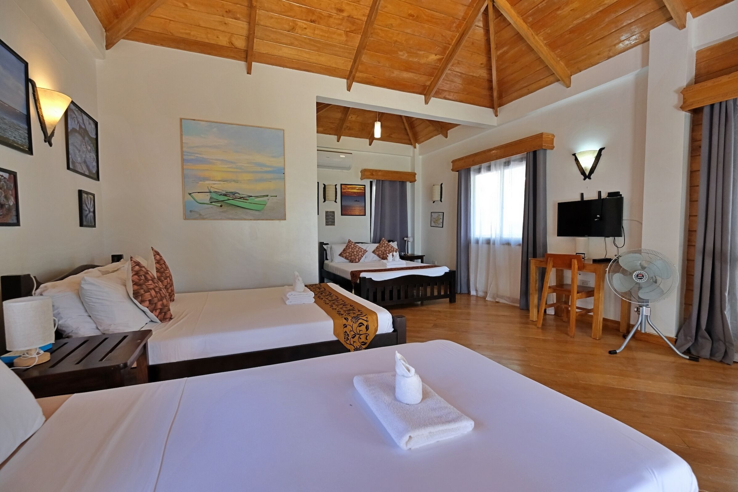 A hotel room with two beds, a wooden ceiling, a painting, a TV, and a fan.