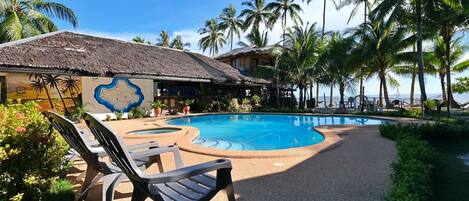 A pool area with lounge chairs, a thatched-roof building, and palm trees.