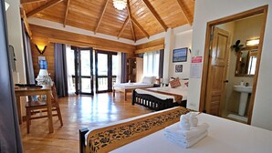 Een hotelkamer met een houten plafond, een bed met een bloemenbedekking, een bureau met een stoel, een groot raam en een badkamer die door een open deur zichtbaar is.