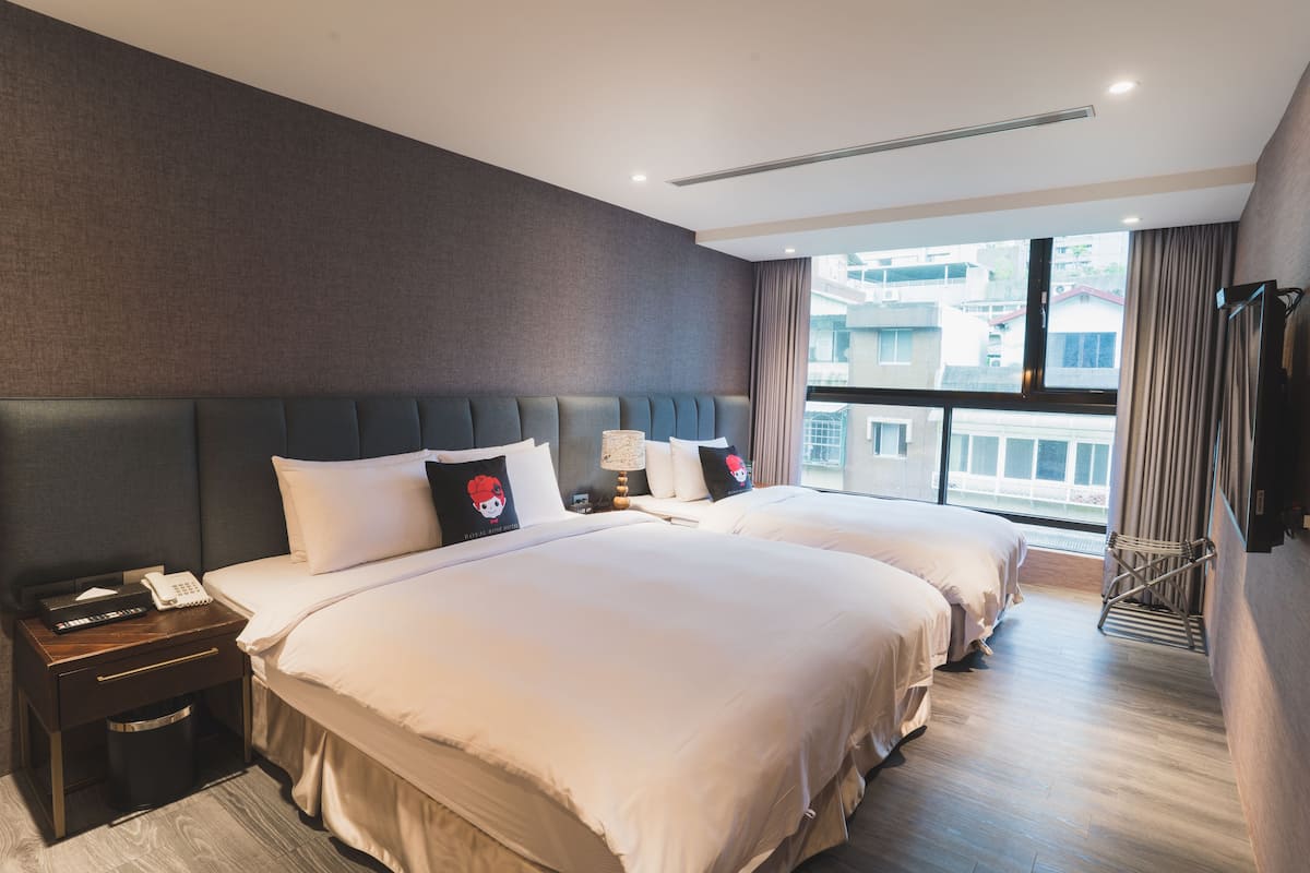 Habitación de hotel con dos camas, una mesita de noche con teléfono y lámpara, y vistas a edificios a través de amplias ventanas.