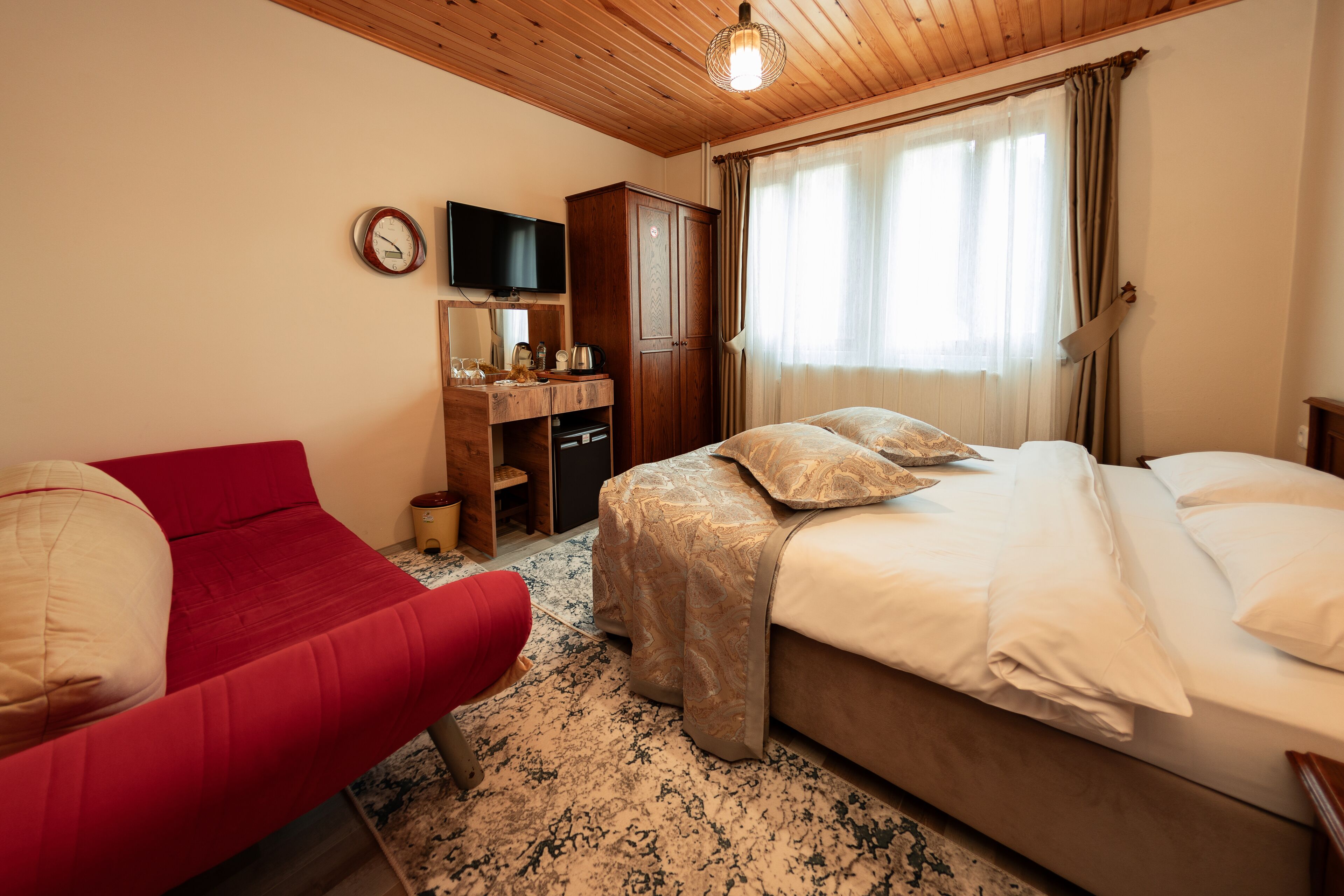 A hotel room with a bed, a red sofa, a wooden wardrobe, a television, and a window with curtains.