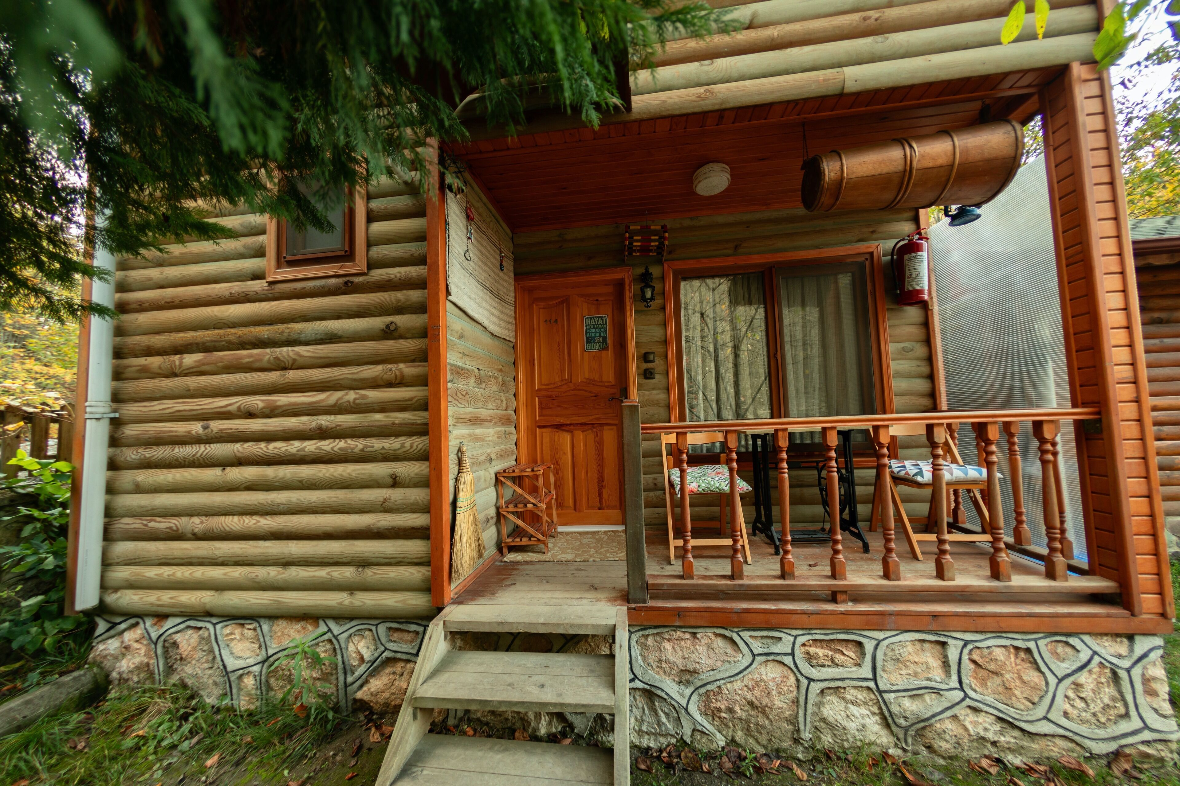 A log cabin with a stone foundation, wooden steps leading to a porch, and a fire extinguisher mounted on the wall.
