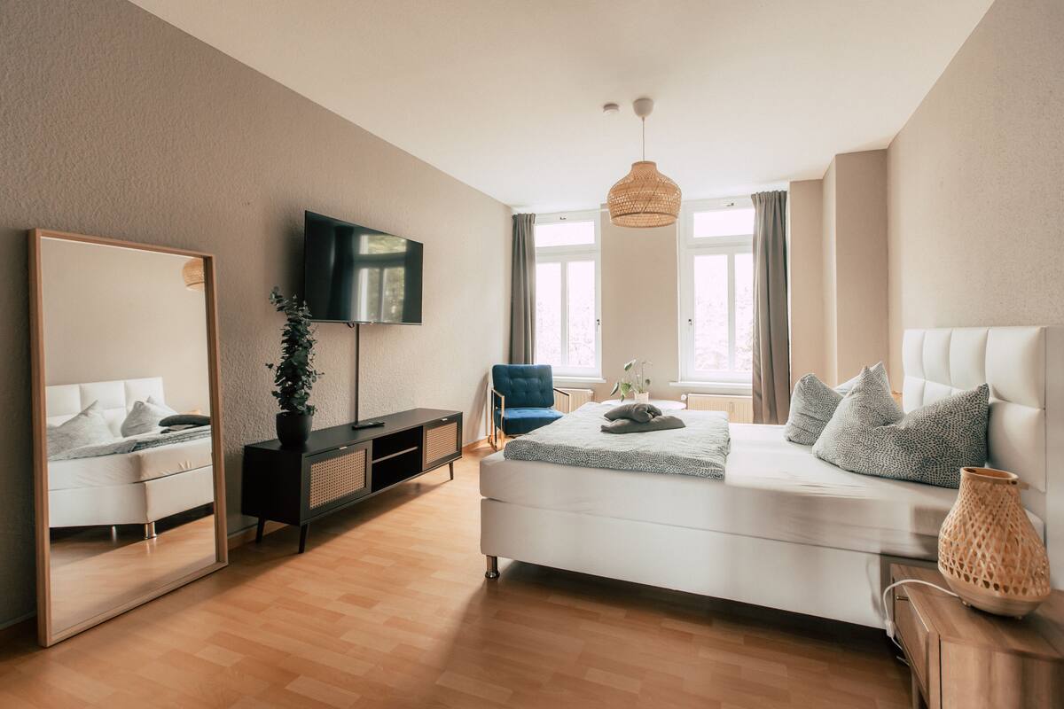 A modern bedroom with a large bed, a TV, a mirror, and a plant.