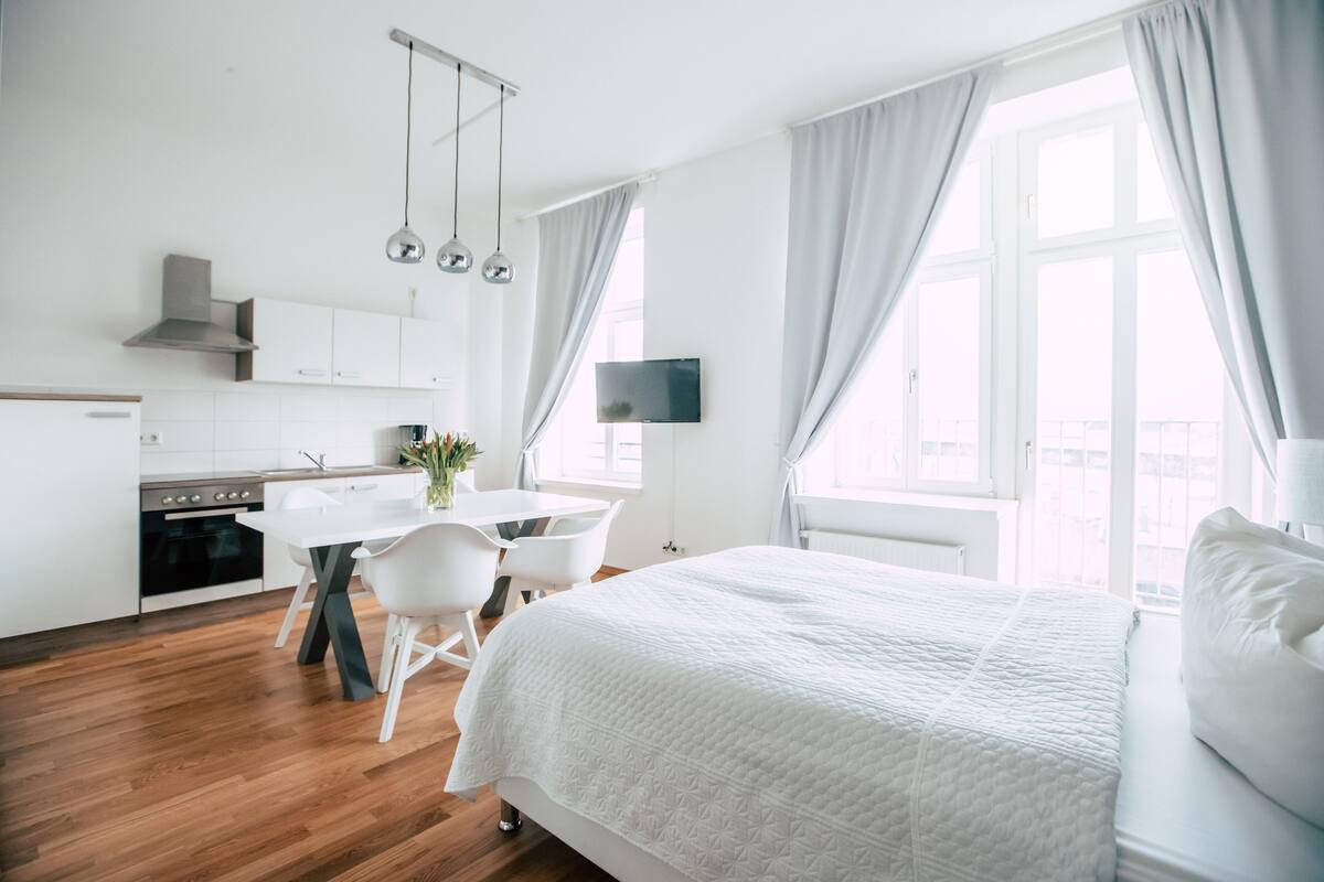 A modern hotel room with a large bed, a dining table with chairs, a kitchen area, and large windows with curtains.