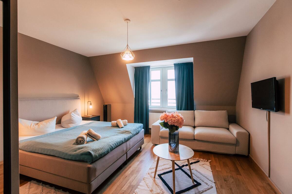 A modern hotel room with a bed, a sofa, a TV, and a window with curtains.