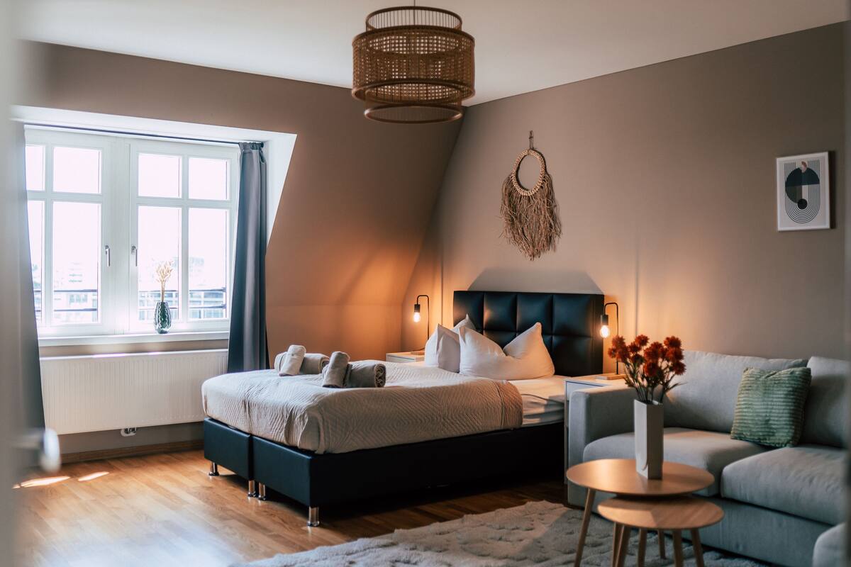 A bedroom with a bed, a bedside table, a lamp, a window, and a view of the city.
