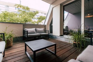 Terraza de madera con un sofá y una mesa, rodeada de plantas en macetas.