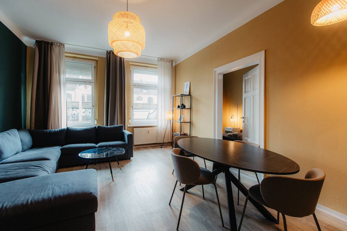 A modern living room with a sofa, a round table, and chairs. There are two large windows with curtains, a standing lamp, and a shelf with books.