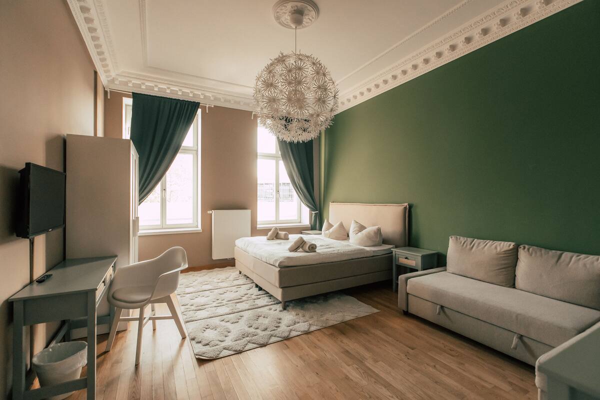 A bedroom with a bed, a desk, a chair, a sofa, and a chandelier.
