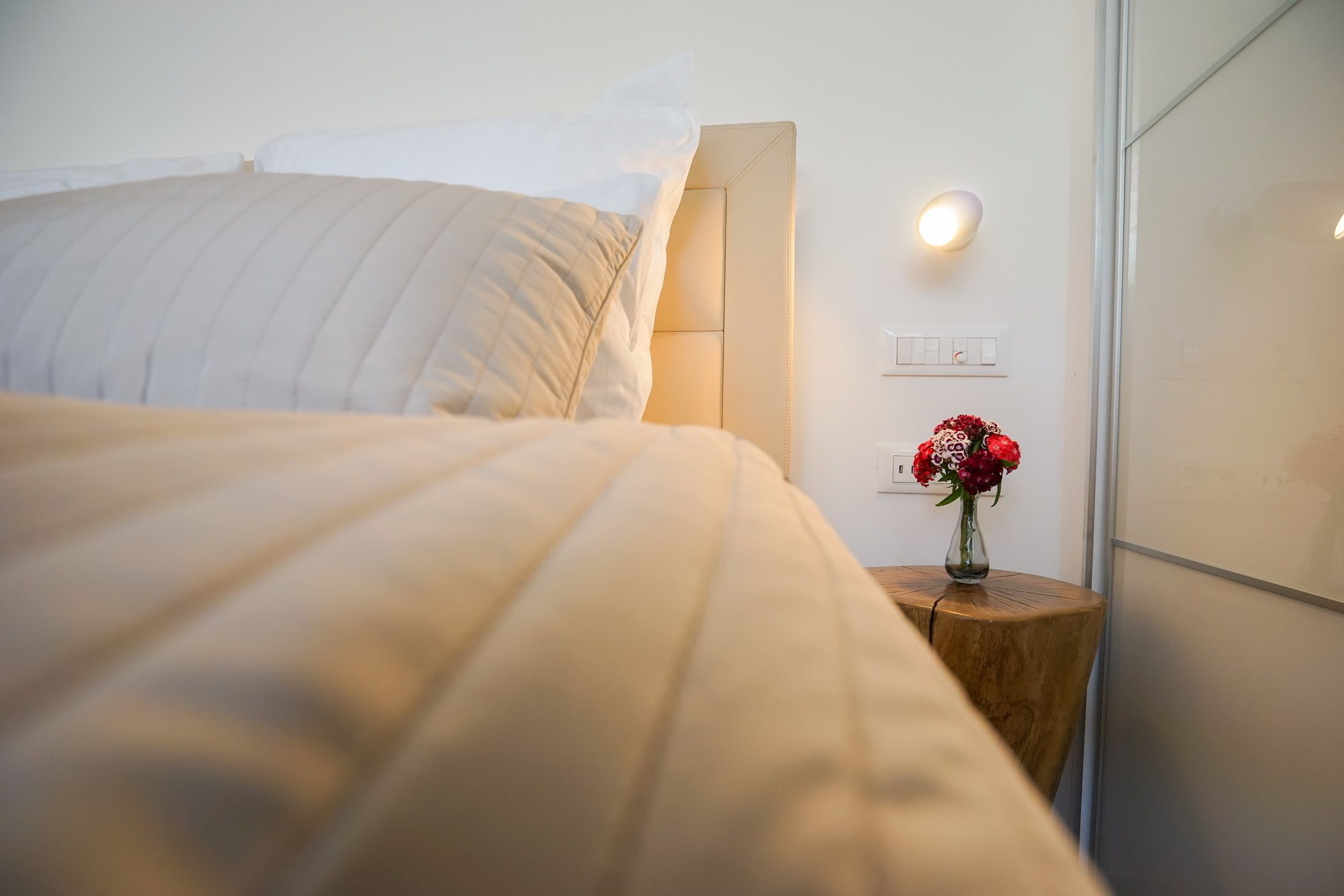 Ein ordentlich bezogenes Bett mit Kopfteil, ein Nachttisch mit Blumenvase und eine Wandleuchte.