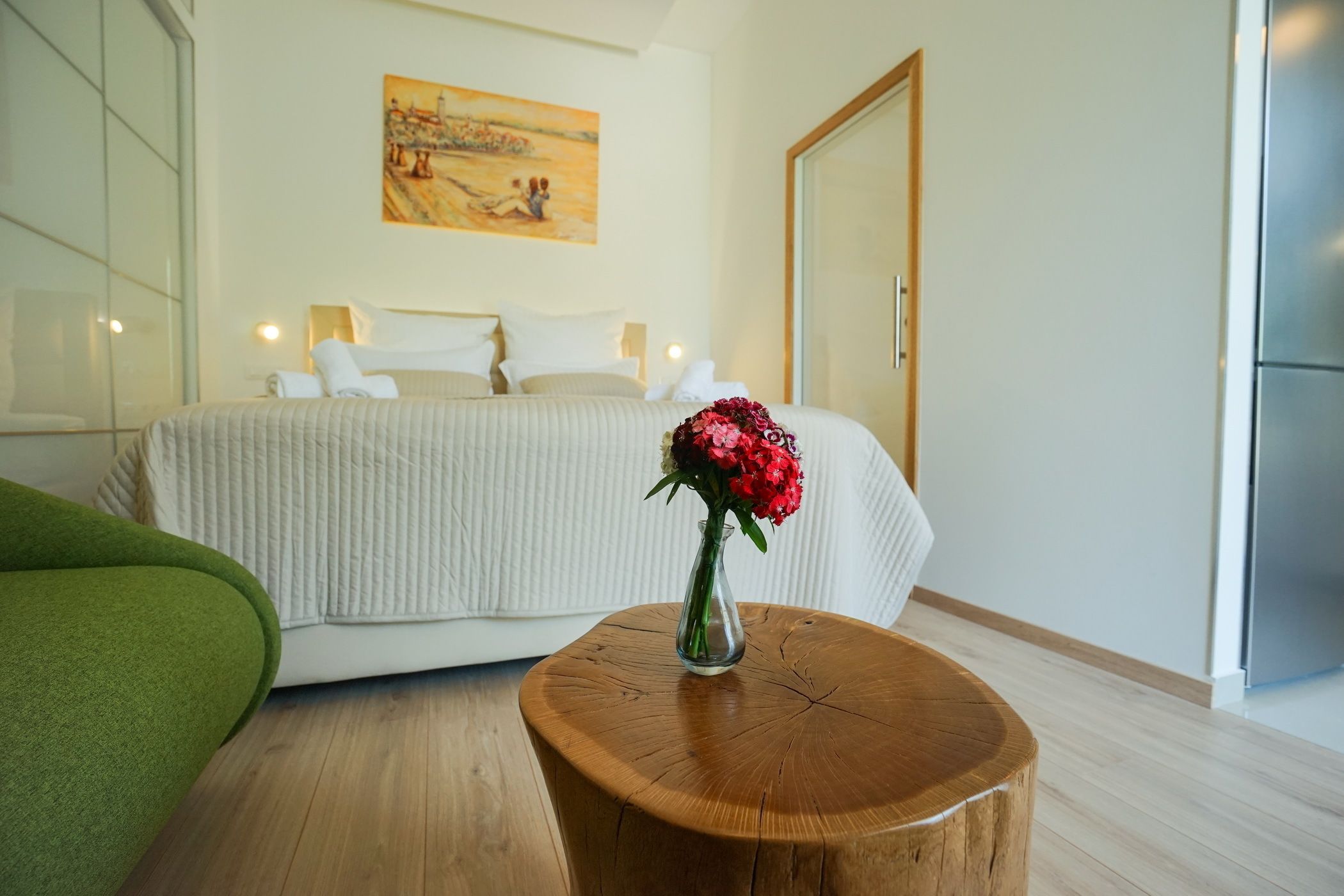 Habitación de hotel con cama, una mesa redonda de madera con un florero y flores, un sillón verde y un cuadro en la pared.