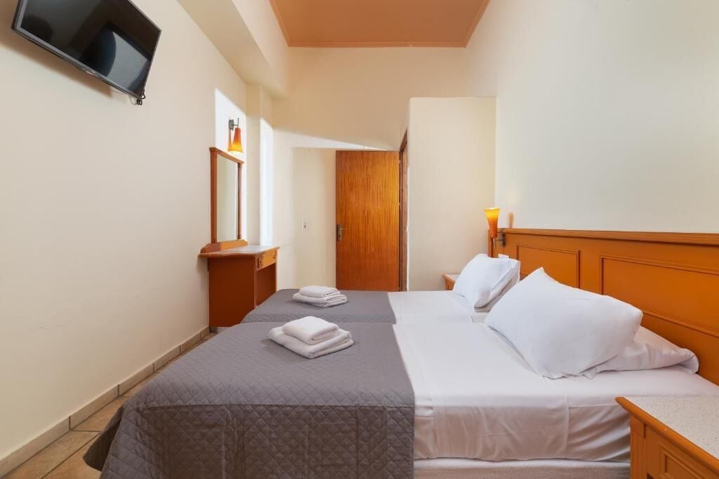 A hotel room with two beds, a TV mounted on the wall, a wooden door, and a bedside table with a lamp.