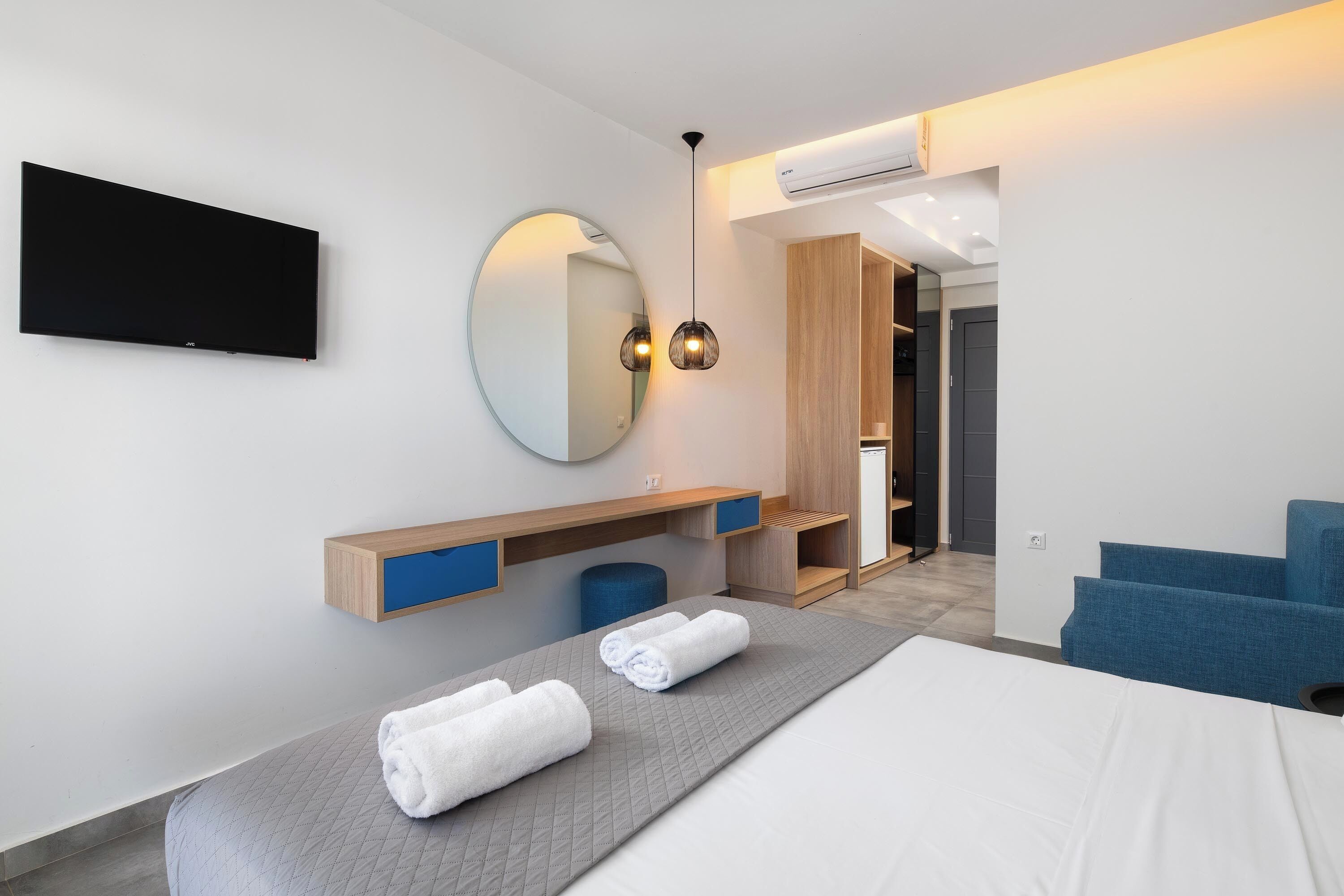 A modern hotel room with a bed, a flat-screen TV, a round mirror, a wooden vanity, and a blue chair.