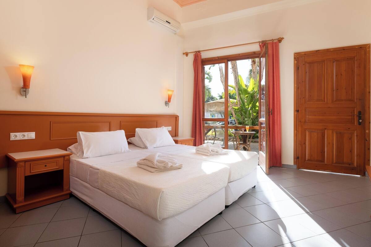 A hotel room with two beds, a wooden nightstand, a window with curtains, and a door.